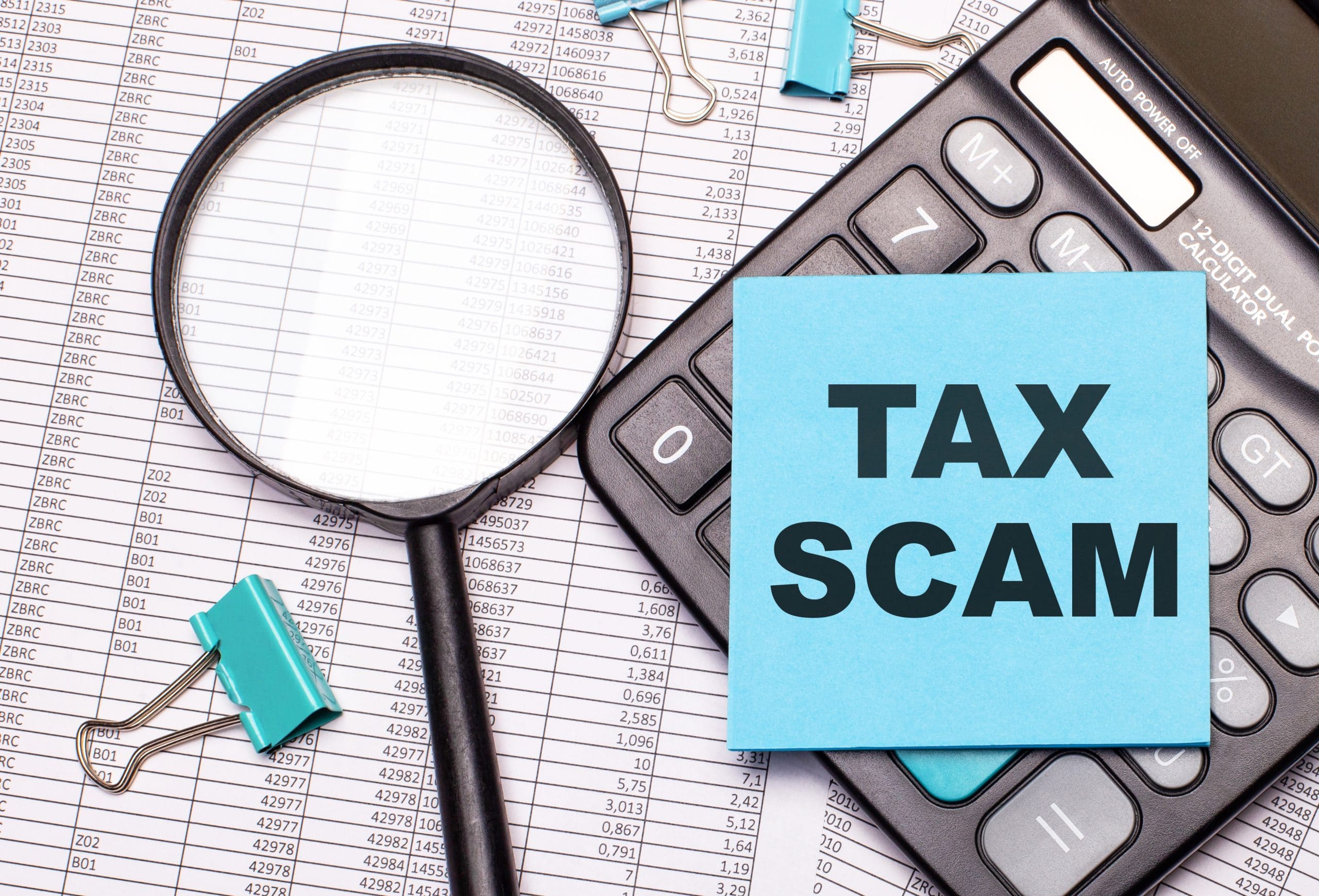 Blue sticky note reading "TAX SCAM" on a calculator, with a magnifying glass and spreadsheets beneath.