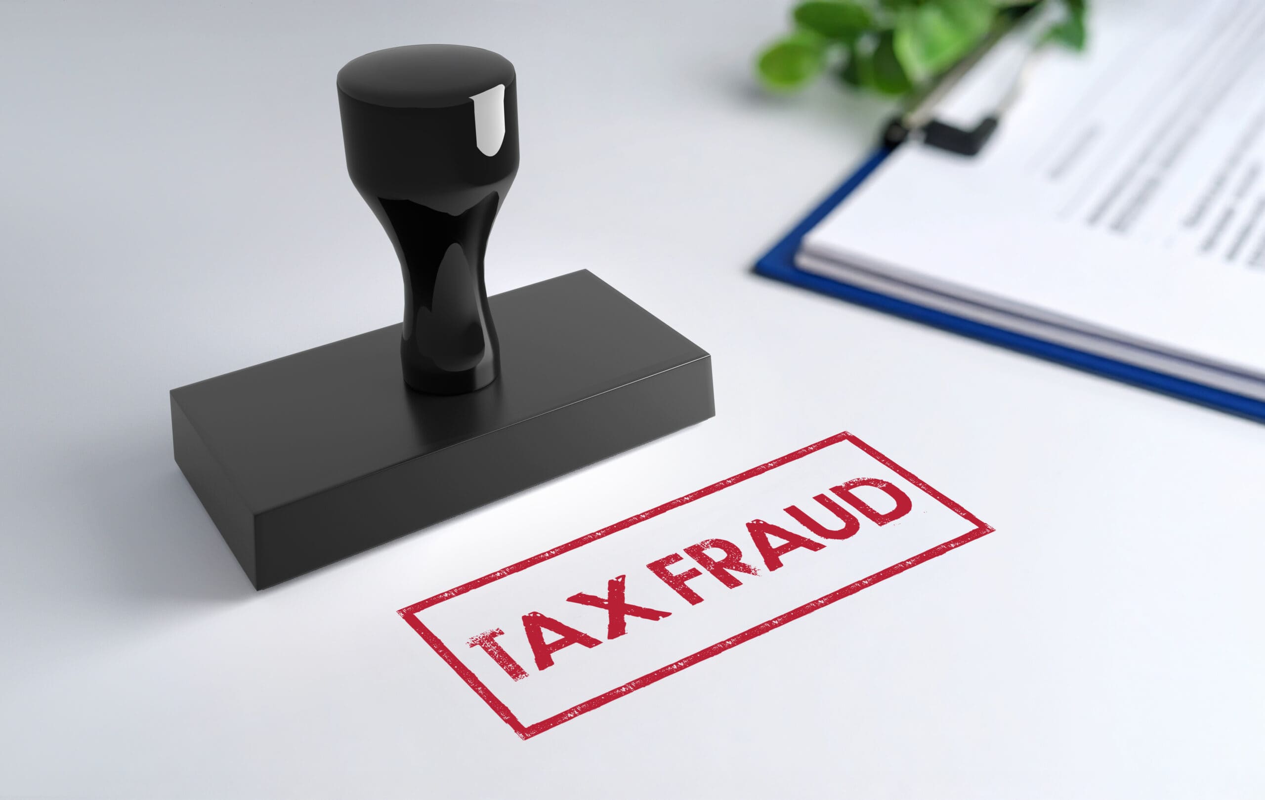 Black rubber stamp sits beside a red "TAX FRAUD" imprint on white paper atop a desk, with a clipboard blurred nearby.