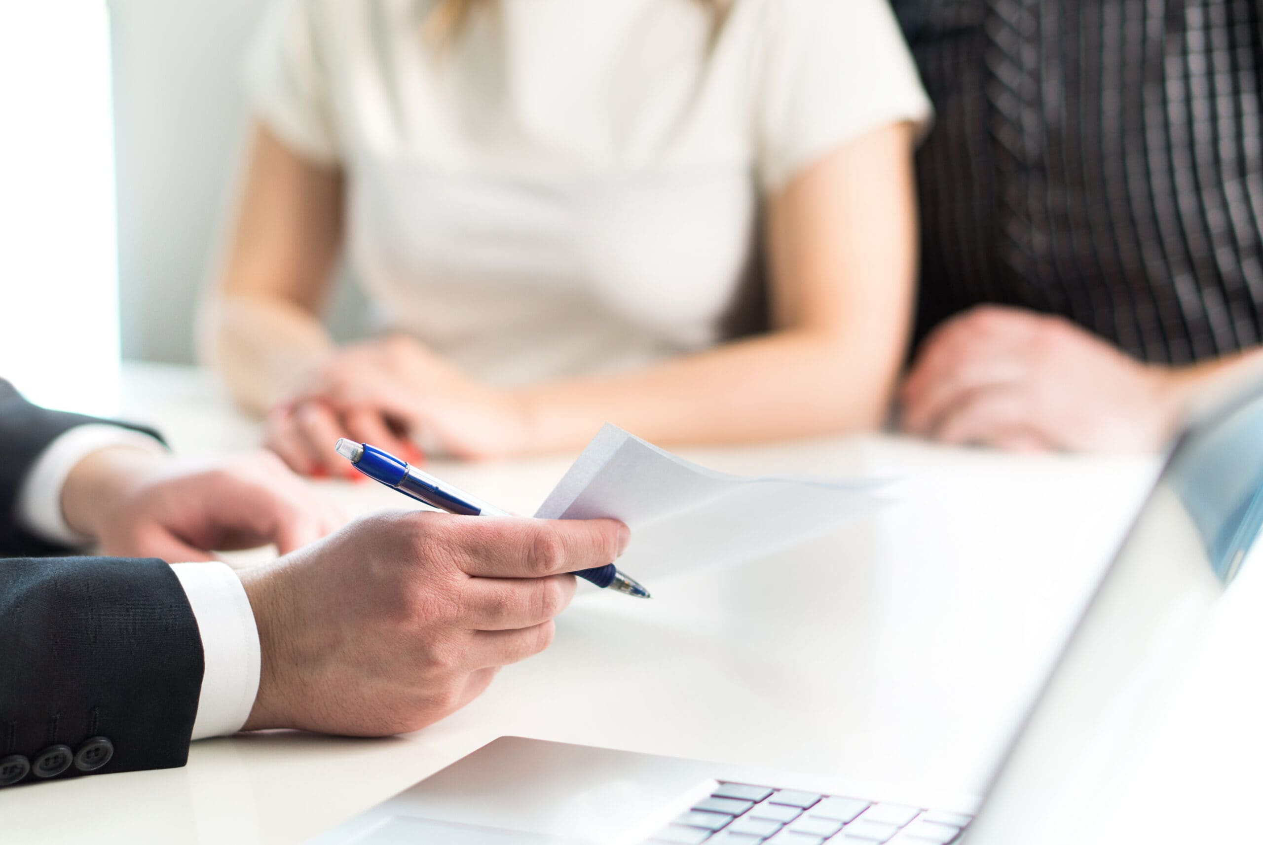 Hand holding a pen over paperwork, poised to sign, at a bright desk with blurred people and a laptop.