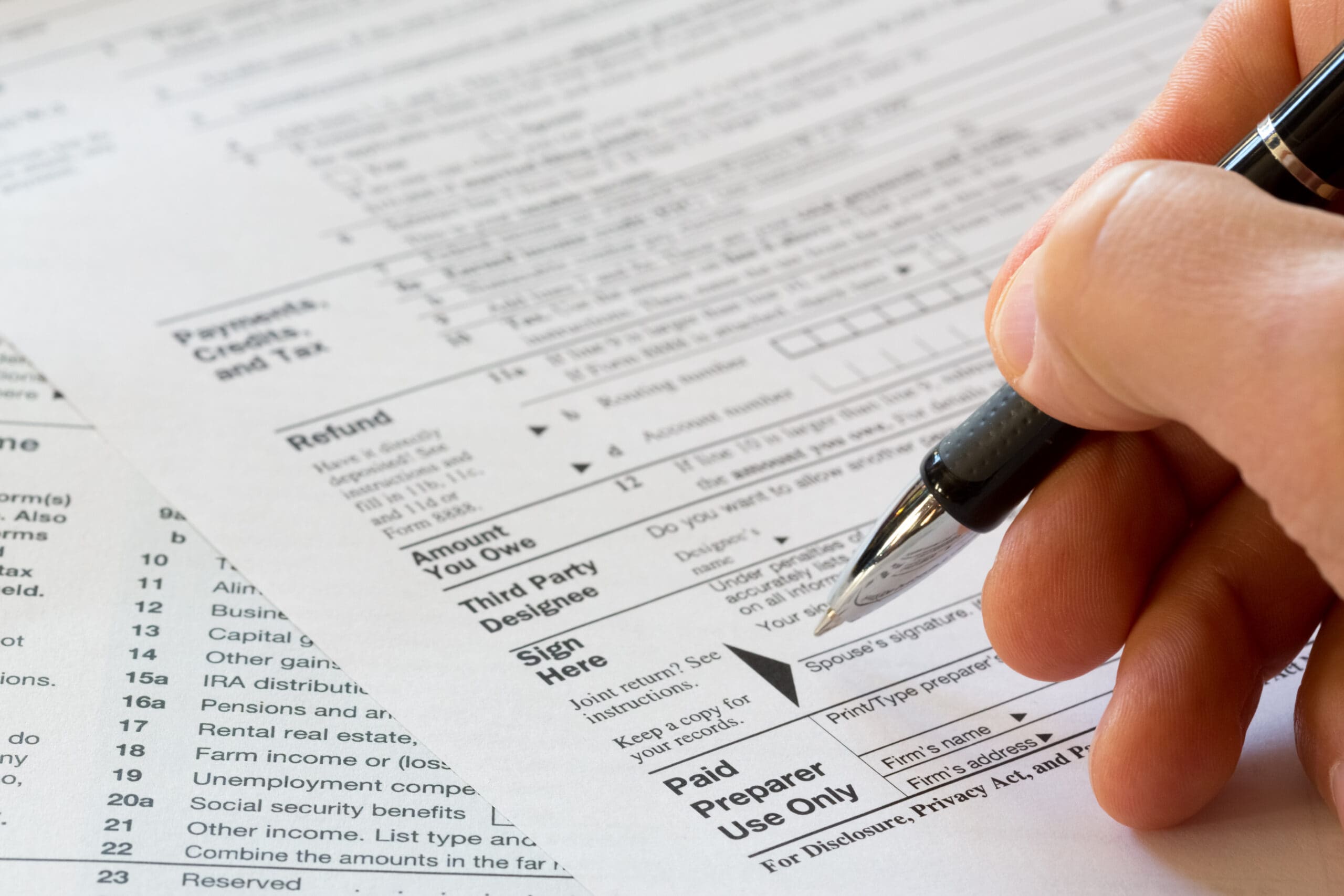 Hand grips a pen poised to sign a tax form resting on printed financial documents.