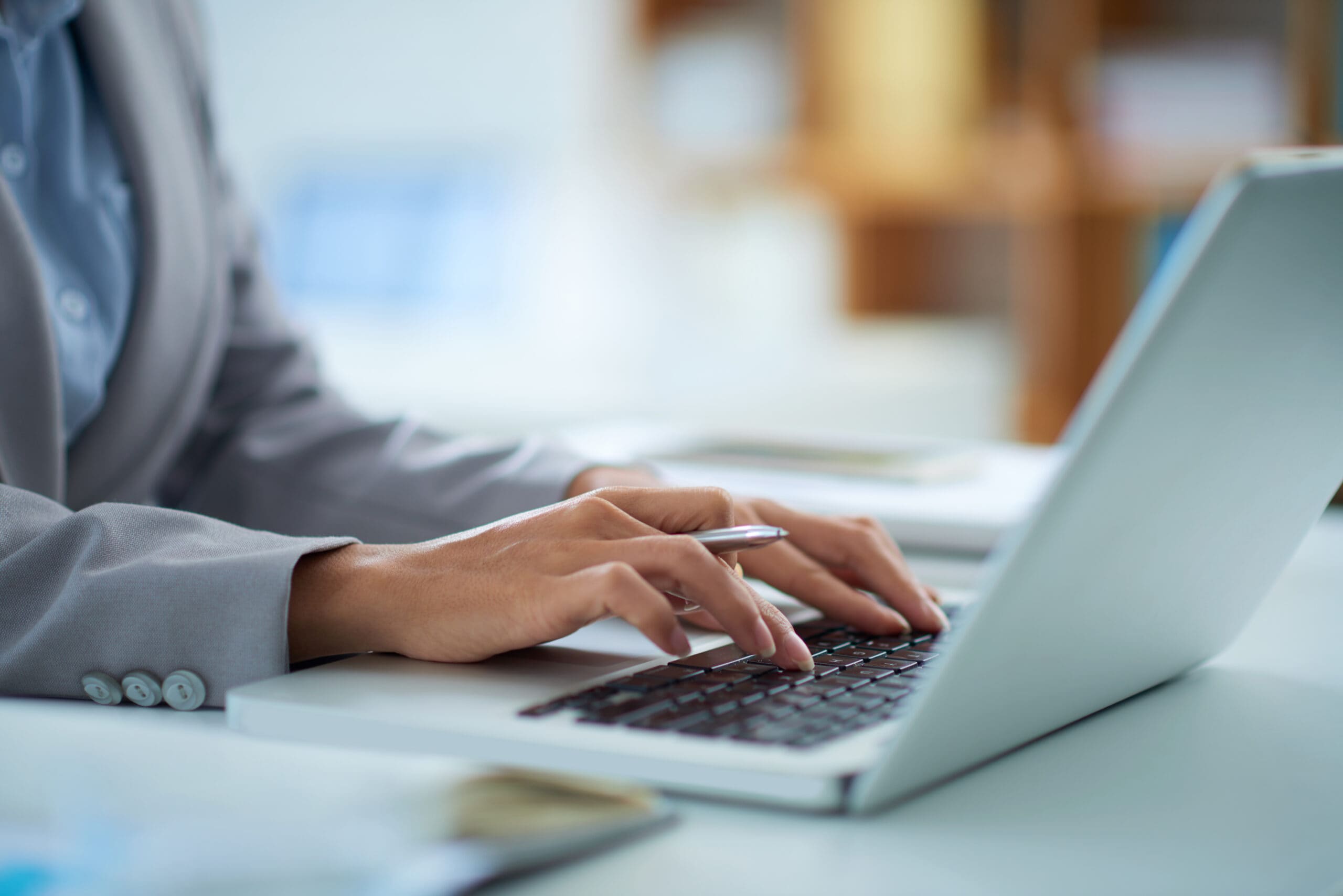 Hands typing on a laptop keyboard, one holding a pen, at a bright desk in an office.